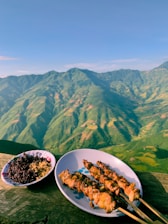A plate of skewered grilled meat sits on a wooden surface, accompanied by a bowl of dark rice and seasoning. In the background, lush green mountains extend under a clear blue sky, providing a picturesque view.