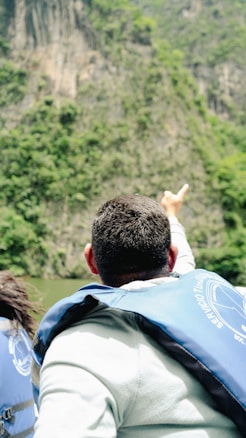 A person wearing a blue life jacket points towards a steep, lush green cliff. The setting suggests a natural landscape with water possibly in the background.