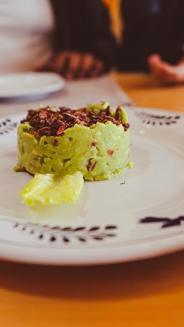 A plate features a neatly shaped serving of guacamole, topped with crispy insect pieces, likely crickets. A small piece of tortilla chip is placed beside the serving. Two hands and a blurred figure in the background suggest a dining setting.