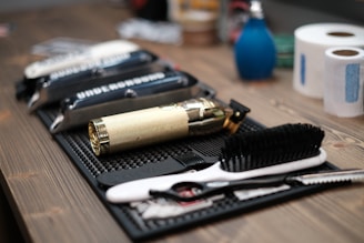 A neatly arranged set of barber tools on a wooden surface, including a gold-colored electric hair clipper, several combs, scissors, and a black brush. In the background, there are two rolls of white tissue and a blue bottle.