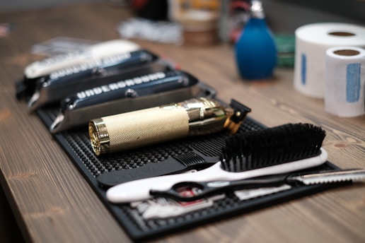 A neatly arranged set of barber tools on a wooden surface, including a gold-colored electric hair clipper, several combs, scissors, and a black brush. In the background, there are two rolls of white tissue and a blue bottle.