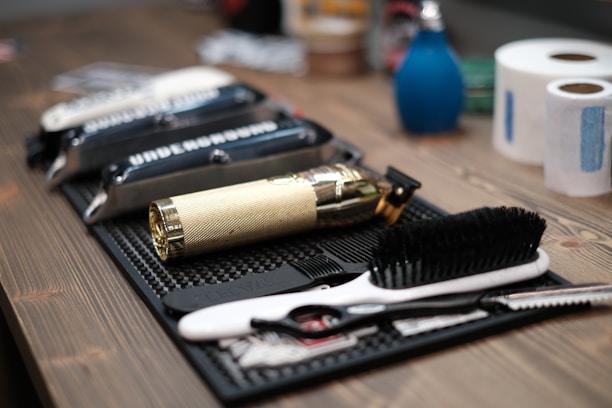 A neatly arranged set of barber tools on a wooden surface, including a gold-colored electric hair clipper, several combs, scissors, and a black brush. In the background, there are two rolls of white tissue and a blue bottle.