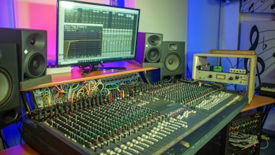 A professional audio production studio setup featuring a large mixing console with numerous knobs and sliders in the foreground. A computer monitor in the background displays audio mixing software with visible equalizer settings. Surrounding the monitor are several large speakers, indicating a focus on sound engineering and clarity. Various cables and audio equipment are organized across the surfaces, contributing to a busy and technical atmosphere. The lighting in the room is a combination of blue and pink hues, creating a dynamic and modern ambiance.