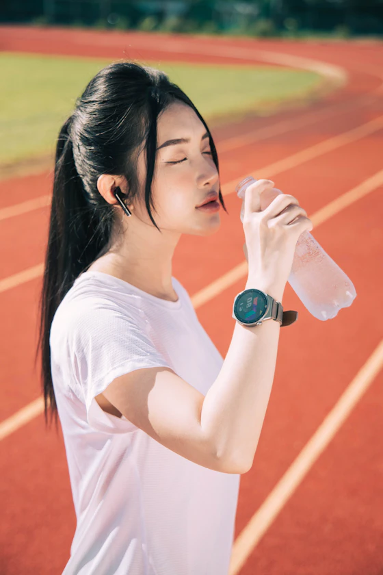 Corredora de camisa branca em pista de atletismo bebendo água, com relógio esportivo exibindo dados no pulso