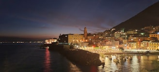 A coastal town is illuminated under a twilight sky, with a prominent clock tower standing amidst a cluster of colorful buildings. The reflection of lights shimmers on the calm sea, and several small boats are docked in a marina. The surrounding hills are faintly visible against the darkening sky.