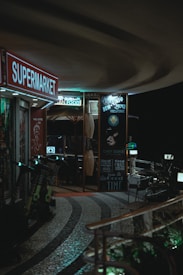 A dimly lit walkway features a supermarket sign and an array of neon lights advertising various services and shops. Scooters are parked along the sidewalk by a restaurant, which has chairs and tables set up outside. The atmosphere is intimate and cozy, with a curved pathway leading through the scene.