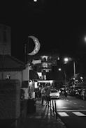 A nighttime urban street scene with a sign shaped like a crescent moon displaying the word 'LOCAL', and a neon 'BAR' sign nearby. People are gathered on the sidewalk, and cars are parked along the dimly lit street. A 'RENT A CAR' sign is visible in the background, with a modern building's glass facade softly illuminated.