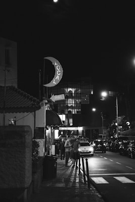 A nighttime urban street scene with a sign shaped like a crescent moon displaying the word 'LOCAL', and a neon 'BAR' sign nearby. People are gathered on the sidewalk, and cars are parked along the dimly lit street. A 'RENT A CAR' sign is visible in the background, with a modern building's glass facade softly illuminated.