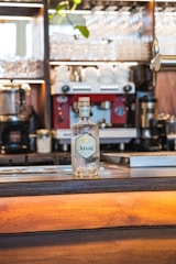 A clear glass bottle labeled 'Anaë' is placed on a wooden bar counter. In the background, a red espresso machine is surrounded by glassware and a few green leaves hanging from above. The setting appears to be a bar or café with a warm, ambient light.