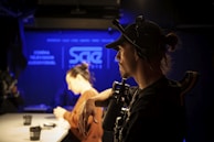 A person wearing a cap and headset is operating a camera in a dimly lit studio with blue accents. Another individual in the background is blurred, sitting at a table with cups and equipment.