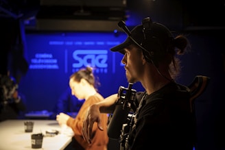 A person wearing a cap and headset is operating a camera in a dimly lit studio with blue accents. Another individual in the background is blurred, sitting at a table with cups and equipment.