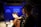 A person wearing a cap and headset is operating a camera in a dimly lit studio with blue accents. Another individual in the background is blurred, sitting at a table with cups and equipment.