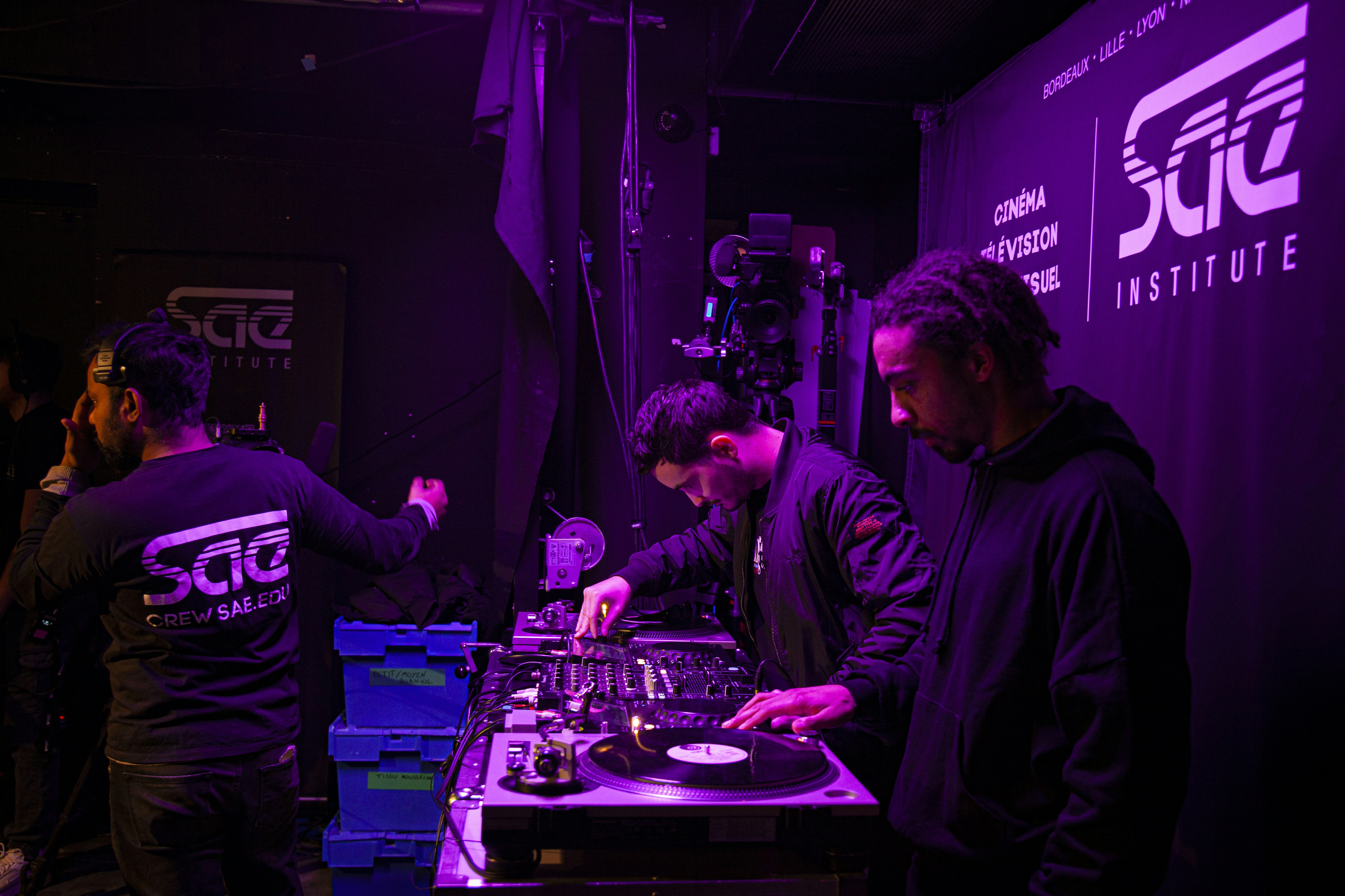 A group of people are engaged in a music production or DJ session in a dimly lit room. Two individuals are focused on turntables and audio equipment, possibly preparing or playing music. The setting features a backdrop with the SAE Institute logo and several blue storage boxes are visible on a table, suggesting an organized working environment.
