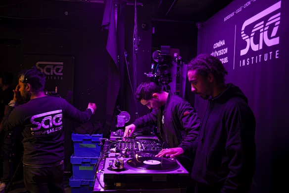 A group of people are engaged in a music production or DJ session in a dimly lit room. Two individuals are focused on turntables and audio equipment, possibly preparing or playing music. The setting features a backdrop with the SAE Institute logo and several blue storage boxes are visible on a table, suggesting an organized working environment.