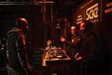Several people are gathered in a dimly lit room with DJ equipment and a camera. Two individuals are behind a DJ booth, one of them pointing at something while wearing sunglasses. Another person is standing nearby, facing them. The walls display signage for an institute involved in cinema and music.