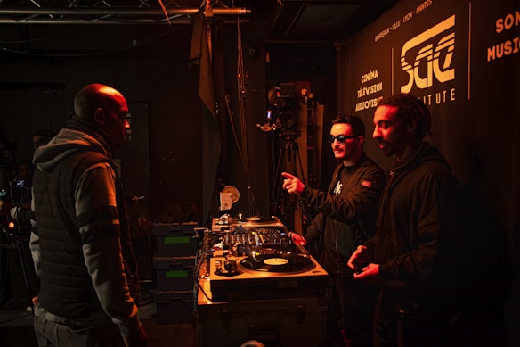 Several people are gathered in a dimly lit room with DJ equipment and a camera. Two individuals are behind a DJ booth, one of them pointing at something while wearing sunglasses. Another person is standing nearby, facing them. The walls display signage for an institute involved in cinema and music.