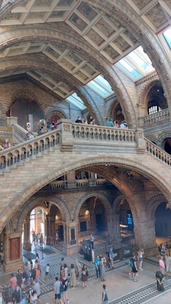 An interior view of a grand, historic building with detailed stone architecture and high arched ceilings. There are intricate patterns on the ceiling and walls and a large, open, multi-level space. People are walking around, exploring the site, which features large windows allowing natural light to illuminate the space.