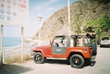 A red Jeep with an open top is parked on a coastal road next to a beach. Nearby, there are steep rocky cliffs with some greenery at the base. A large informational sign is positioned close to the road, along with a streetlamp.