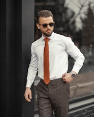 A well-dressed man stands confidently outdoors. He is wearing a white shirt, a striking orange tie, and dark trousers, complemented by sunglasses. His hair is neatly styled, and he has a trimmed beard. The setting appears to be urban with reflective surfaces and muted surroundings.