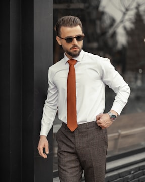 A well-dressed man stands confidently outdoors. He is wearing a white shirt, a striking orange tie, and dark trousers, complemented by sunglasses. His hair is neatly styled, and he has a trimmed beard. The setting appears to be urban with reflective surfaces and muted surroundings.