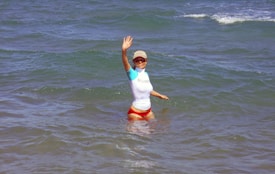 A person is standing in the ocean waves, wearing a white and blue shirt and red shorts. They are waving with one hand and wearing a cap and sunglasses.