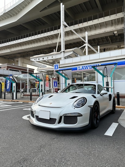 A sleek white sports car is parked in front of a convenience store, set under a large overpass. The store has blue and white signage, and the scene is urban with visible concrete and steel structures. The car's design is aerodynamic and modern, with a low profile and prominent wheel arches.