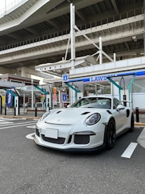 A sleek white sports car is parked in front of a convenience store, set under a large overpass. The store has blue and white signage, and the scene is urban with visible concrete and steel structures. The car's design is aerodynamic and modern, with a low profile and prominent wheel arches.