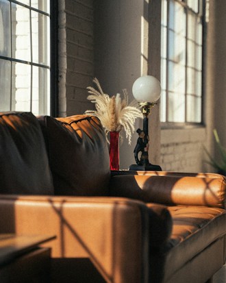 Warm sunlight streams through large windows, casting soft shadows on a tan leather sofa. A red vase with dry pampas grass is placed on a table beside a vintage lamp. The room has a cozy, inviting atmosphere.