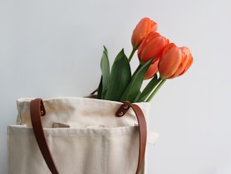 A canvas bag with brown leather straps containing a bouquet of vibrant orange tulips, with green stems and leaves, against a plain white background.