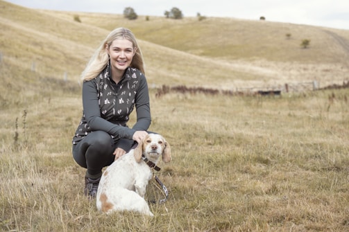 A woman is crouching in a grassy field with rolling hills in the background. She is wearing a dark patterned vest and dark pants, smiling while petting a small white and brown dog. The setting is serene and natural, under a cloudy sky.