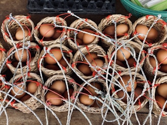 Small woven baskets are filled with brown eggs and arranged in rows. Each basket has a handle made of intertwined rope, some of which have red accents. The baskets are placed on a wooden surface, with another container partially visible in the background.