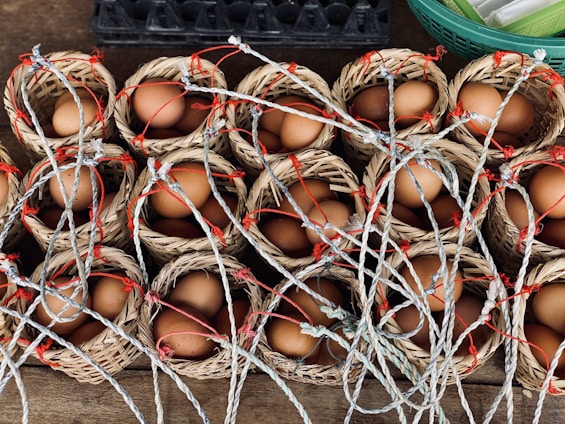 Small woven baskets are filled with brown eggs and arranged in rows. Each basket has a handle made of intertwined rope, some of which have red accents. The baskets are placed on a wooden surface, with another container partially visible in the background.