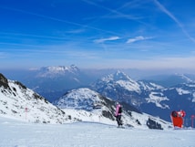 Snow-covered mountains under a clear blue sky create a serene winter landscape. A skier in pink attire is moving down the slope, adding vibrancy to the pristine white surroundings. Red markers along the path indicate ski trail directions, while distant mountain ranges fade into the horizon.