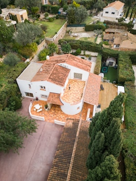 An aerial view of a Mediterranean-style villa surrounded by lush greenery and hedges. The house features red-tiled roofs and a spacious terrace with a smooth, circular pattern. There is a patio area with outdoor furniture and a driveway leading to the residence. The garden is well-maintained with tall trees and decorative plants, providing a serene and private atmosphere.