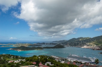A picturesque coastal landscape featuring a vibrant blue sea nestled between lush green islands and mountains. Numerous boats and a large cruise ship are visible in the harbor, creating a bustling yet serene scene. The sky above is partly cloudy, offering dramatic contrasts between the bright areas and shadowed sections.