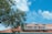 A red-roofed building features a sign reading 'Port of Sale' with the word 'Sale' creatively incorporating a dollar symbol. A large tree with green leaves frames the left side of the image, and a bright blue sky with scattered clouds forms the background.