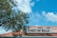 A red-roofed building features a sign reading 'Port of Sale' with the word 'Sale' creatively incorporating a dollar symbol. A large tree with green leaves frames the left side of the image, and a bright blue sky with scattered clouds forms the background.