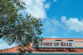 A red-roofed building features a sign reading 'Port of Sale' with the word 'Sale' creatively incorporating a dollar symbol. A large tree with green leaves frames the left side of the image, and a bright blue sky with scattered clouds forms the background.