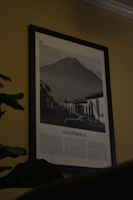 A framed black and white photograph depicts a scene with a prominent volcano in the background. In the foreground, there's a street lined with old buildings and a car parked alongside. Below the photograph, text describes Guatemala.