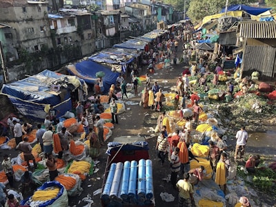 An outdoor market scene bustling with activity. Vendors display vibrant yellow and orange floral garlands on the ground. The area is filled with people engaged in various activities like bargaining and buying. Shops with blue tarpaulin roofs line the street, and the surroundings have a lived-in, rustic appearance.