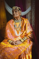 An elderly person dressed in traditional attire sits with a composed expression. The outfit is a brightly colored sari adorned with intricate patterns and paired with ornate jewelry including a headdress, necklaces, and bangles. The background features fabric in various colors, adding to the richness of the scene.