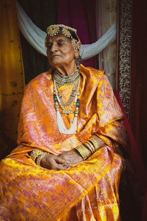 An elderly person dressed in traditional attire sits with a composed expression. The outfit is a brightly colored sari adorned with intricate patterns and paired with ornate jewelry including a headdress, necklaces, and bangles. The background features fabric in various colors, adding to the richness of the scene.