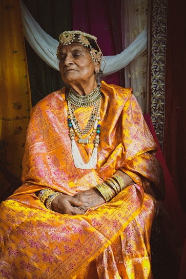 An elderly person dressed in traditional attire sits with a composed expression. The outfit is a brightly colored sari adorned with intricate patterns and paired with ornate jewelry including a headdress, necklaces, and bangles. The background features fabric in various colors, adding to the richness of the scene.