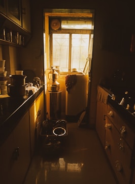 A kitchen illuminated by warm, golden sunlight coming through a window. Stainless steel utensils and containers are arranged on the counters and shelves. Shadows create a cozy and nostalgic ambiance.