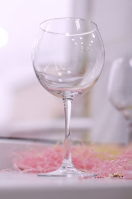 A clear, empty wine glass with a long stem placed against a softly blurred background. The glass reflects light, creating subtle highlights. Below the glass, there is a layer of pastel-colored, crinkled paper, adding a delicate and playful touch.