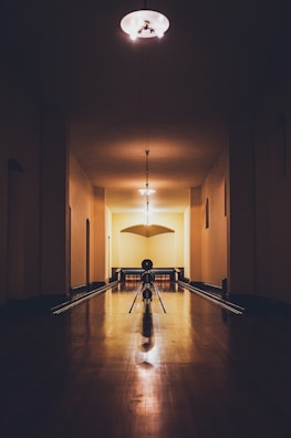 A dimly lit, vintage-style bowling alley with wooden lanes stretches into the distance. The lighting casts warm shadows, illuminating the smooth wooden floor and creating an intimate atmosphere. Bowling pins are lined up at the end of the lane, and a solitary bowling ball rests ready to be rolled.