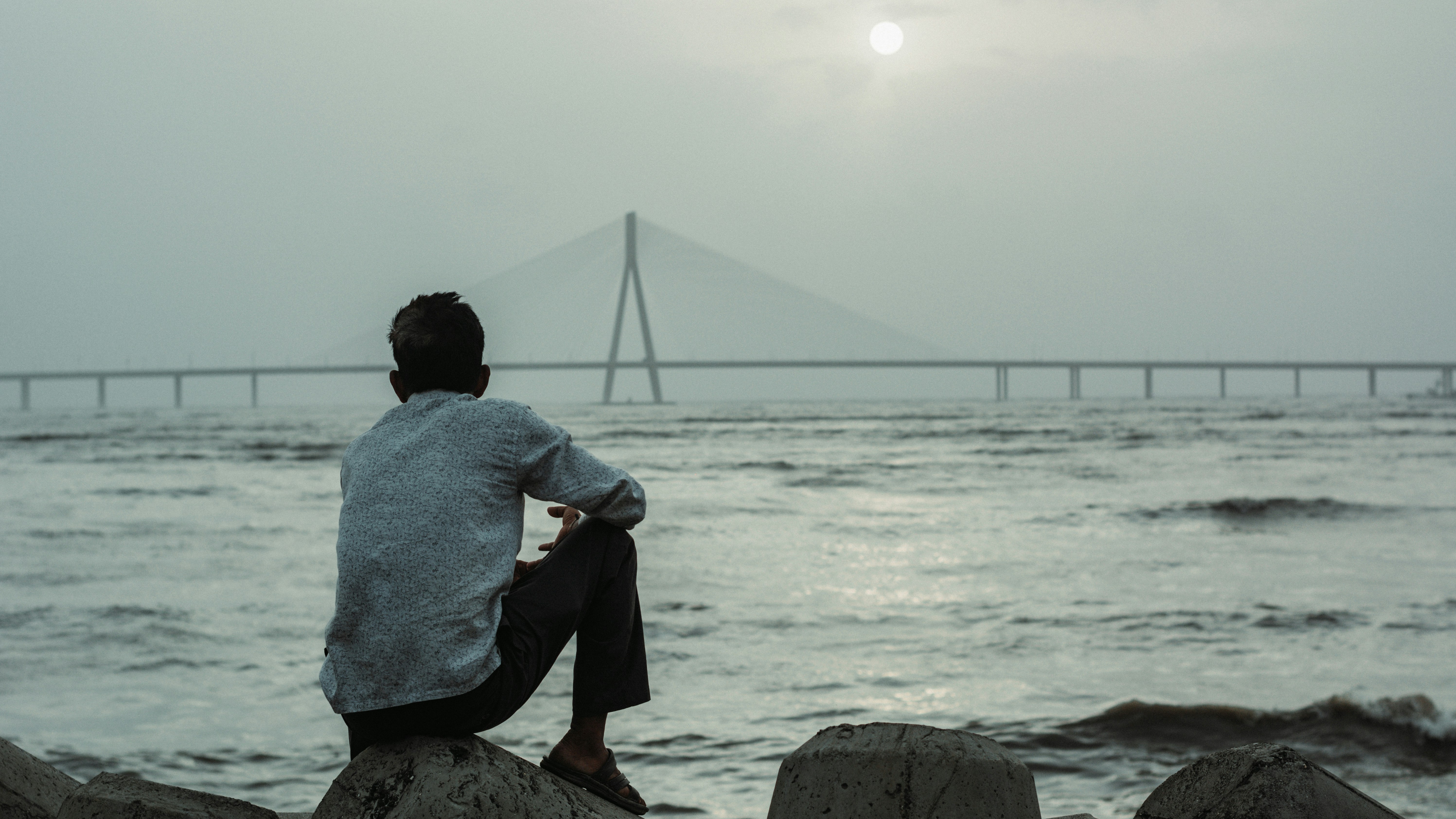 man sitting on the shore staring at a bridge across the sea