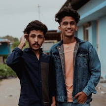 Two young men stand close together, both wearing casual clothing. One wears a dark shirt and the other a denim jacket over a peach shirt, smiling towards the camera. They appear relaxed and are standing on a street with buildings in the background.