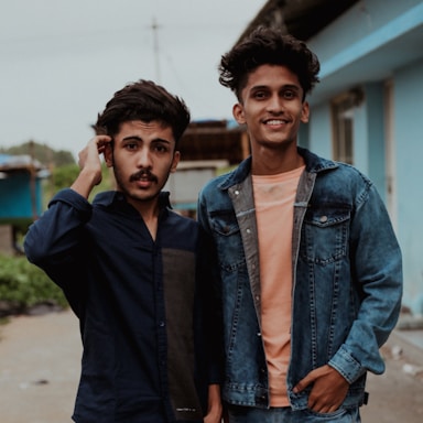 Two young men stand close together, both wearing casual clothing. One wears a dark shirt and the other a denim jacket over a peach shirt, smiling towards the camera. They appear relaxed and are standing on a street with buildings in the background.
