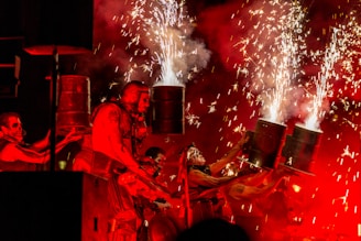A dynamic performance featuring individuals with painted faces using metallic barrels that emit sparks and fireworks. The scene is bathed in intense red lighting, adding a dramatic flair to the energetic display.